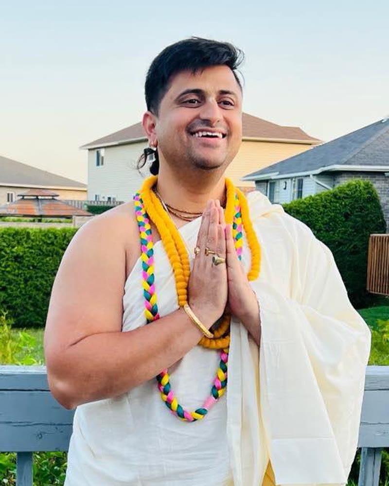 Darshan Upadhyay (Damodar Charan Das) smiling with folded hands in greeting, wearing flower garlands